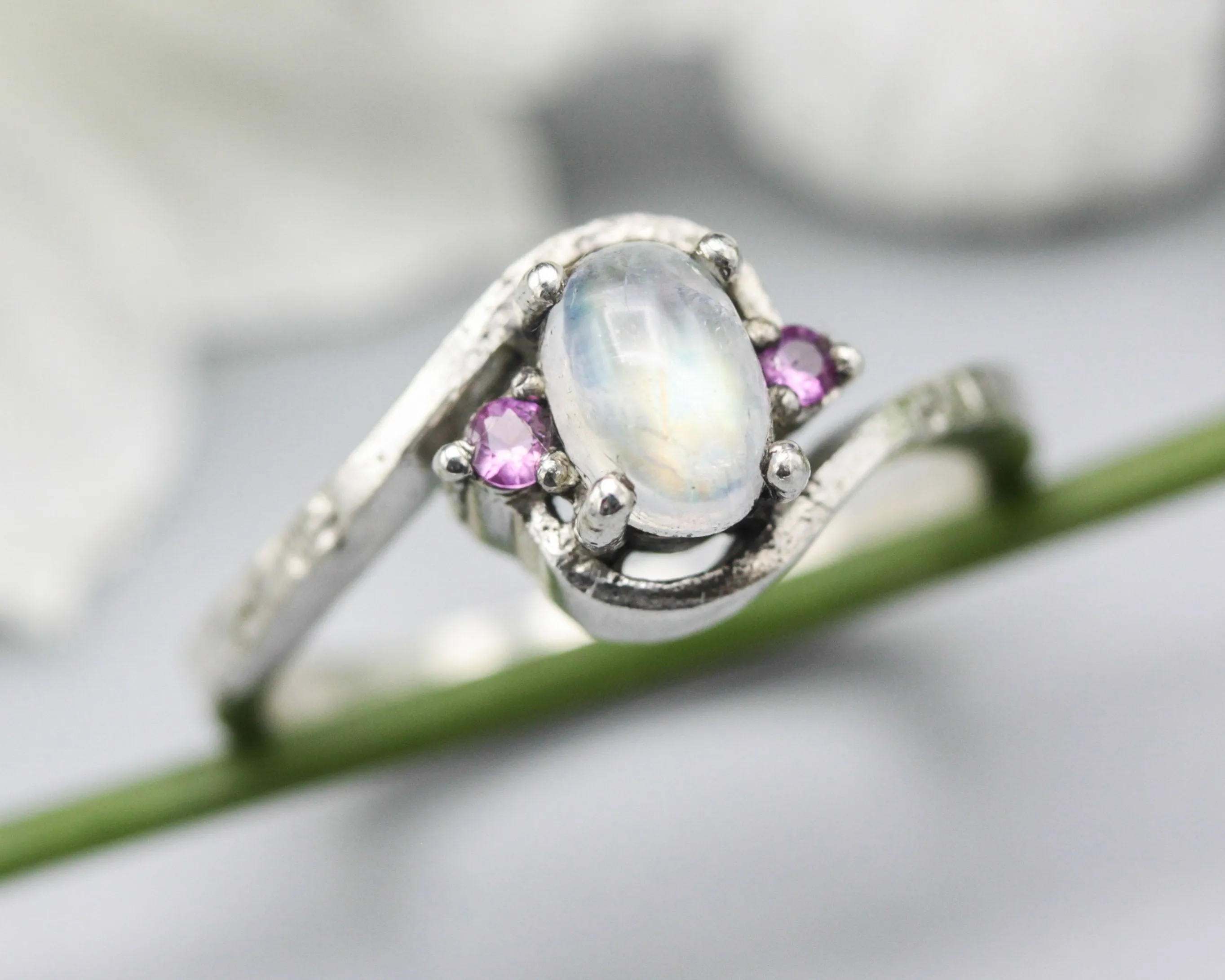 Oval cabochon moonstone ring with pink sapphire side set gems in prongs setting with sterling silver oxidized texture band