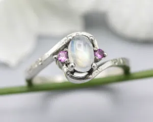 Oval cabochon moonstone ring with pink sapphire side set gems in prongs setting with sterling silver oxidized texture band